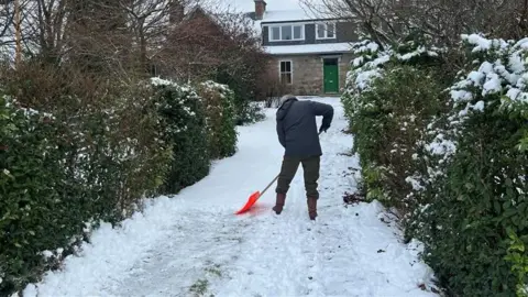 A man, seen from behind, with a snow shovel and standing in his driveway as he shovels snow. His house can be seen in the distance behind him, with a green door.