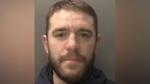 Billy Ormiston - a custody image of a man with short brown hair, a moustache and short beard. He is wearing a dark blue hoody and stood against a grey background