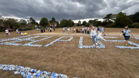 Lucy Barron-Reid Bottles of water being used to spell "Help Us" by residents of Benenden affected by an ongoing outage. There is a border of water bottles alongside the sign. Beneath the sign, the grass is dry and yellow after the summer heatwaves and dry weather. The sky above is dark with rain clouds.
