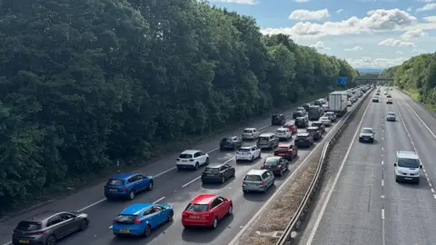 Motorway traffic at a standstill. Stationary cars in all three lanes of motorway.  Cars are moving along the carriageway on the opposite side. The motorway is surrounded by trees. 