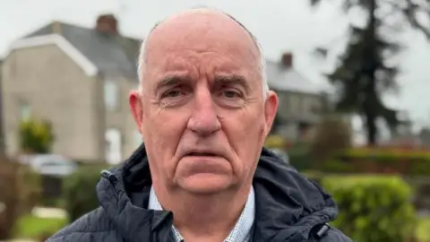 Cllr Trevor Wilson is looking at the camera. He is slightly bald with grey white hair.