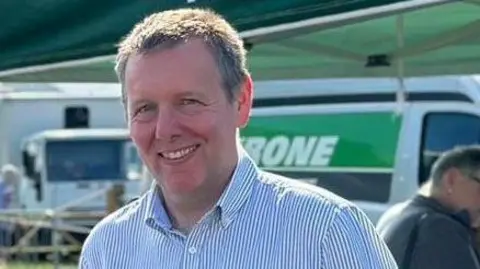 NFU An image of Adam Briggs of the NFU. He has short greying hair and is smiling. He wears an open-neck blue and white shirt.