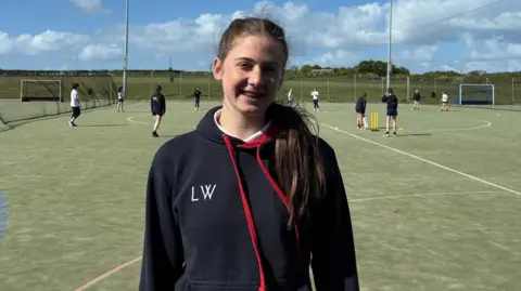 Lily, a pupil at Cockermouth School, stands in front of the astro turf cricket pitch wearing a sports hoodie.