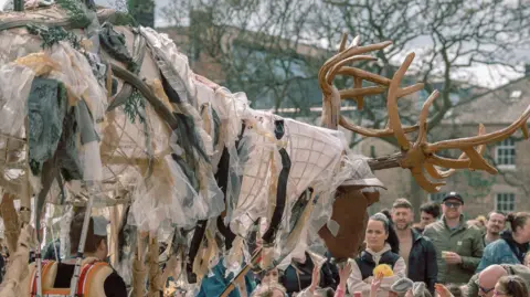 Von Fox Promotions A large puppet of a stag. The body is made out of scraps of material and it has large wooden horns. There are children's hands reaching up to touch the puppet and adults watching on to the right. Georgian buildings can be seen in the background.