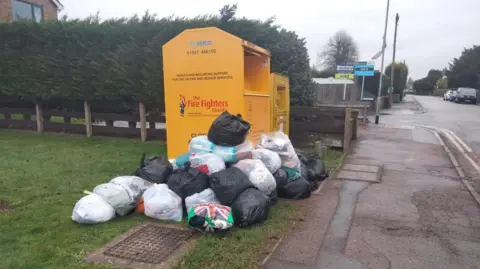 Diane Green Two large yellow metal collection bins for clothes and shoes sit on a piece of grass at a fire station, the bins have lots of bin bags stacked at the front and side, that can not fit it, due to them being full. Some parked cars can be seen on the right.