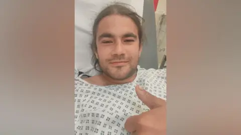 A man in hospital gown and bed, he has long dark hair