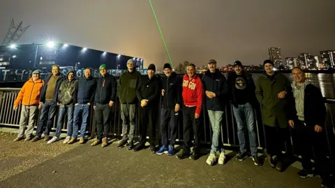 Scott Johnson A group of men on a bridge looking at the camera. They wear warm clothing and the sky is dark.