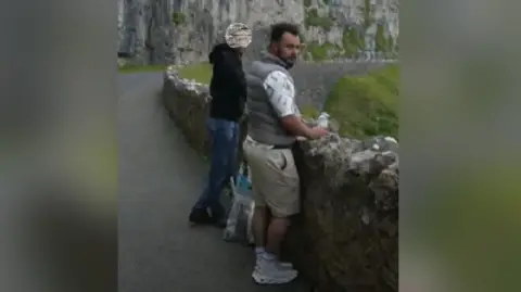 North Wales Police Photo of two men, one close to the camera wearing a grey gillet with beige shorts stood at a rocked wall