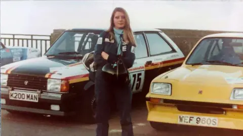 FAMILY HANDOUT Joanna standing in front of two rally cars in the 1980s. The one on the left is black with a yellow and red stripe. The one on the right is yellow.