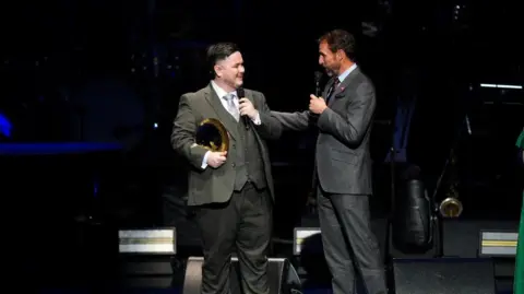 PA Media Award-winner Brandon, who is dressed in a light grey suit holds a golden plate-style trophy, as Southgate in a darker grey suit appears to pat him on the back on stage as he congratulates him