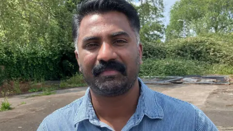 Eldhose Mathew has black hair and black facial hair, both of which are greying. He's wearing a blue and white stripy shirt and is smiling at the camera. There are trees behind him.