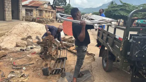 Metal scrap dealers at work alongside a road in the center of Moroguro. A man holds a car door shell moving to his position on the back of a truck. Another person behind him carry two pieces of metal on the floor.