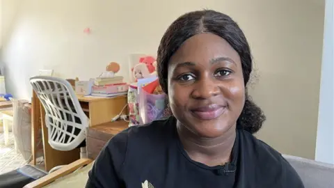 Kelechi Ajiero in a black t-shirt, sat on a sofa and smiling.