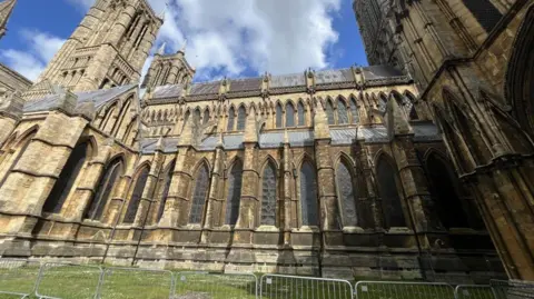The south side of Lincoln Cathedral which has grey railings at the front of a patch of grass in front of the grand building. Several long pointy windows rise next to pillars at the side of the building.