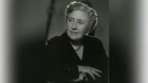 An archive sepia image of Agatha Christie, around 1950. She is sitting down with her arms folded and looking away from the camera.