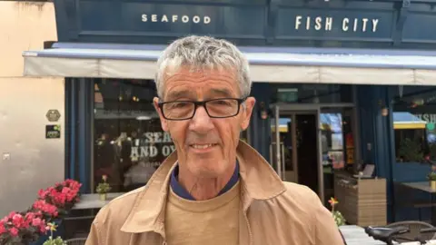 John Lavery, a man with grey hair and glasses, in a brown jacket, outside the premises of Fish City in Belfast.