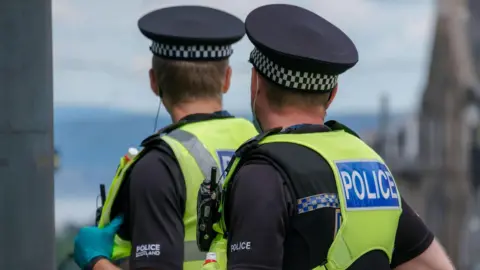 The back of two police officers heads. They are both wearing black hats and hi vis Police vests.