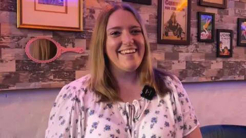 A woman with light brown shoulder length hair smiles at the camera. She's wearing a white blouse with blue flowers, sitting in front of a wall of mirrors and picture frames.