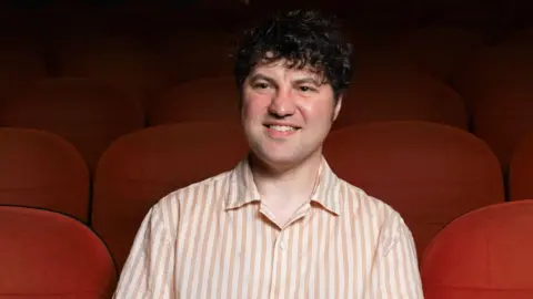 Ryan Rutherford Paul Gallagher - a man with dark hair and a beige coloured shirt - sits in a seat in a cinema