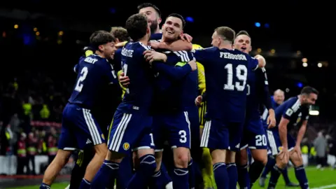 PA Media Several Scotland footballers embrace on the pitch. They are wearing an all blue strip with white trim.