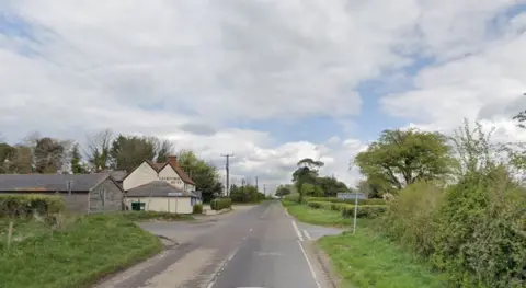 Road junction looking with a small lane running left to right there are road signs on the left side.  To the left is a barn and other buildings.