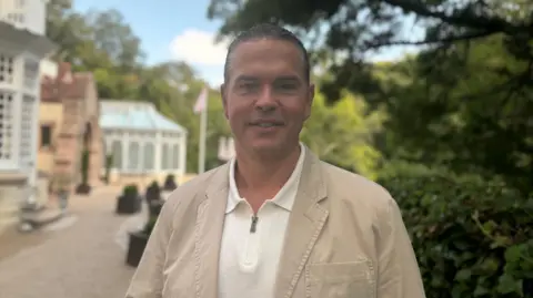Marcus Calvani is pictured in a white polo shirt and beige jacket. He has short black hair and is smiling. The gardens of Chateau la Chaire are visible behind him. Part of the granite building is also visible.