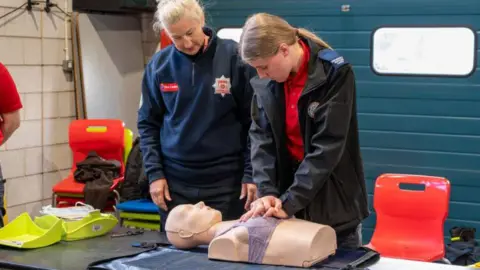 Northamptonshire Police, Fire and Crime Commissioner Two women, standing over a female CPR dummy. There is a red chair to the left, and equipment around it. The women are in dark outfits. 