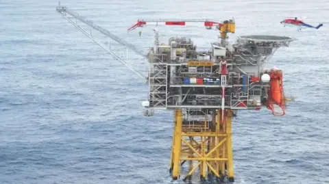 Harbour Energy Oil and gas platform with yellow legs in the sea, a blue, red and white offshore helicopter hovers overhead, and vessels in the background. 