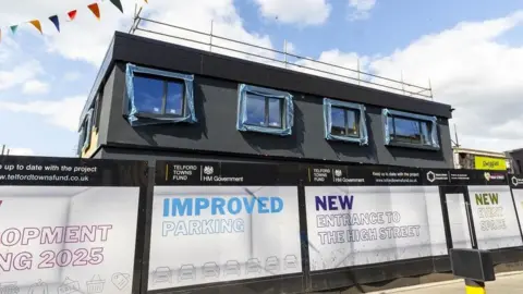 Telford and Wrekin Council A black flat-roof building behind barriers with the UK government logo and Telford Towns Fund logo on it. The unit has scaffolding on the top and has blue plastic over the window frames.