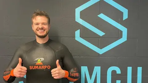Stuart Hacker Tony Hopkin in a back wetsuit with his thumbs up. He is smiling.