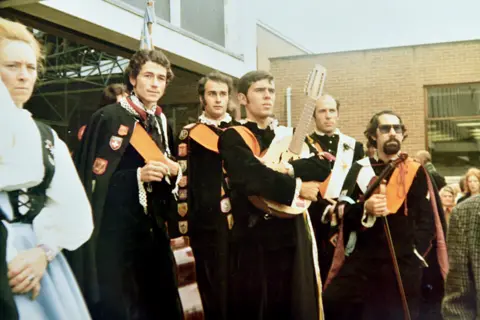 Walter Crinnion A group of men wearing black cloaks. One man in middle is holding a 12-string guitar. The man on the right hand side is wearing sunglasses and has a thick goatee beard. There is a woman on the left of the photo in green and white traditional dress.