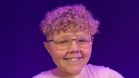 Curley haired boy with glasses and smiling at camera with white t-shirt.