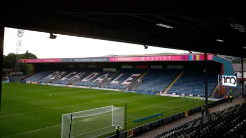 A view from behind the goal at Rochdale's Crown Oil Arena, with a view of the stand along the touchline with 'ROCHDALE' painted across the seats. 