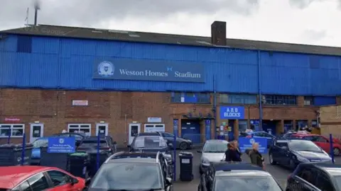 Google The exterior of Peterborough United FC's stadium, which features a blue corrogated panel on the top half of the building while the rest is made up of red bricks. There are cars parked up in two rows in front.
