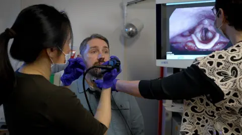 A man sits in a chair as his doctor and speech therapist view images of his vocal cords on a screen
