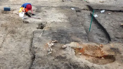 Cotswold Archaeology An archaeologist crouched down over a shallow trench with their left gloved hand resting on the earth and their right gloved hand hovering above the trench. 