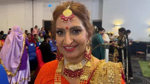 A woman with ginger hair and a red dress, wearing gold necklaces, earrings and head decoration.