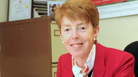 PA Media A woman seated at a desk in an office environment, positioned close to the camera. She is wearing a bright red jacket, a patterned scarf, and pearl earrings, with shelves and folders visible in the background. The setting appears professional, with documents and framed items arranged behind the desk. The lighting is even and warm, highlighting the subject and creating a clear, focused portrait.