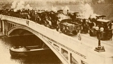 A black and white concrete bridge with one person standing on it next to many steam-powered vehicles all parked on it. 