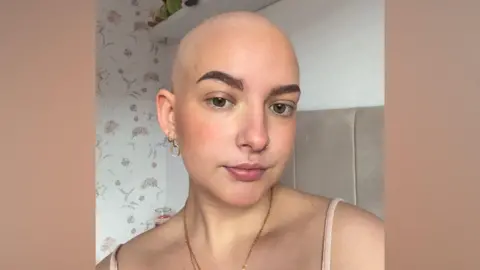 Lilli Hales A woman with no hair is taking a selfie with a serious expression. She has bold brown eyebrows and is wearing a gold necklace, vest, and dainty hooped earrings. Behind her is a light brown headboard and a flower patterned wallpaper.