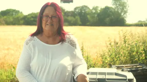 A woman with red hair wearing a white T-shirt standing in front of a field. The tops of bins are to the right of the frame.