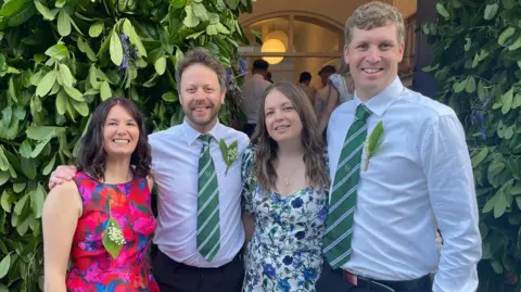 Two couples with their arms around eachother smiling at the camera. The men are wearing white shirts with green and white-striped ties and the women are wearing floral dresses. They are each wearing a sprig of Lily in the Valley - the women's are pinned upside down while the men's are upright. There is green foliage hanging to their left and right and an open door behind them through which are three men wearing white shirts.