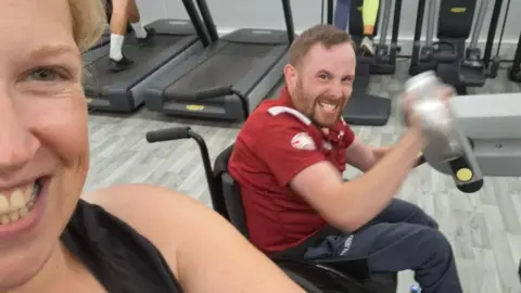 Jordan Burns Two people are in a gym with treadmills in the background. One person in the foreground is smiling toward the camera, while the other is seated in a wheelchair, wearing a red sports shirt and lifting a silver dumbbell. The floor has a gray wood-like finish.