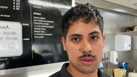 Tarun Singh, 21, has short dark hair and a small matching moustache, with some stubble on his chin. He is wearing a black T-shirt and behind him is the price list at his chip shop.