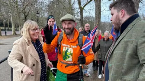 A man in a flat cap with an orange hoodie and rucksack with two union flags attached surrounded by supporters on a walk