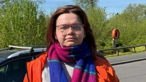 Charlie Stubbs / BBC There is a woman with dark shoulder-length hair, rimless glasses, a blue and purple scarf, a red coat and a light grey jumper. She is stood in front of the road, with a black car over her left shoulder. There is also green trees behind her.