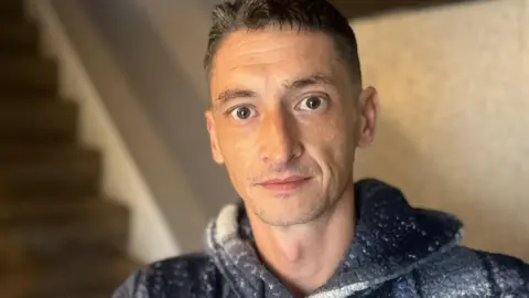 A man with black hair looking at the camera wearing a blue and white jumper. 