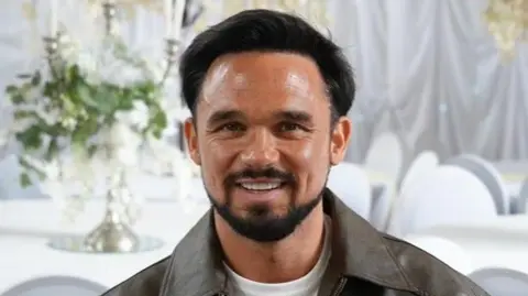 Shaun Whitmore/BBC Head and shoulders image of Gareth Gates in a room with white tablecloths, white chairs and white sheets on the wall. He has short brown hair, beard and moustache, and is wearing a white T-shirt and a brown jacket. 