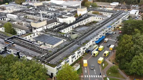 Aerial view of the Queen Elizabeth Hospital in King's Lynn. It is a sprawling grey building surrounded by trees and roads. 