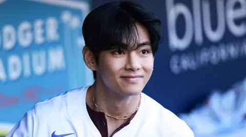 V smiles as he looks at a distance, wearing a baseball shirt